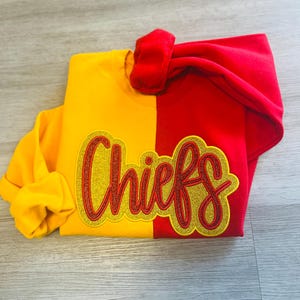 May include: A two-toned sweatshirt, split vertically with yellow and red. The word "Chiefs" is embroidered in a gold glitter script with a red outline. The sleeves are folded, and the hood is visible.
