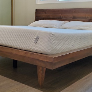 May include: A wooden bed frame with a slatted headboard and a white mattress. The bed frame has a dark brown finish and is raised on four legs.