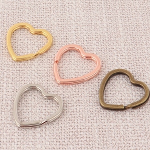 Op de afbeelding: Vier hartvormige sleutelhangers in goud, roségoud, zilver en brons. De sleutelhangers zijn gemaakt van metaal en hebben een eenvoudig, elegant ontwerp. Ze zijn gerangschikt op een getextureerd, lichtgekleurd oppervlak.