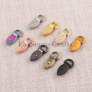 May include: A collection of decorative metal purse clasps in various colors, including silver, gold, rose gold, and black. Each clasp features a teardrop-shaped leatherette base with a metal ring for attachment. The clasps are designed for bags and accessories.