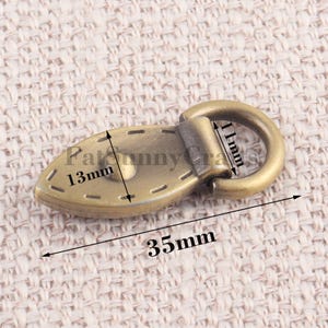 May include: A bronze-colored metal oval-shaped clasp with a ring, measuring 35mm long, 13mm wide, and 11mm in ring diameter. The clasp has decorative stitching around the edges and is likely used for bags or other craft projects.
