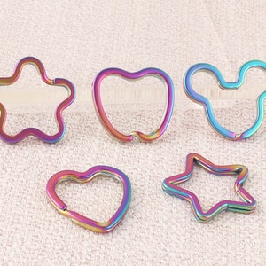 Op de afbeelding: Vijf verschillende sleutelhangers van metaal met een regenboogkleurige, iriserende afwerking. De vormen omvatten een ster, een hart, een appel, een Mickey Mouse-hoofd en een ster.