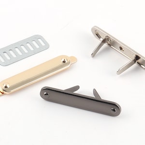 May include: Three metal clasps for bags or purses. One is gold, one is silver, and one is black. The clasps are rectangular and have a small, round knob on each end. The clasps are open and ready to be attached to a bag or purse.