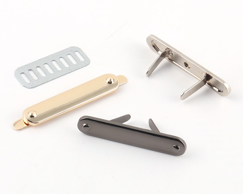May include: Three metal clasps for bags or purses. One is gold, one is silver, and one is black. The clasps are rectangular and have a small, round knob on each end. The clasps are open and ready to be attached to a bag or purse.