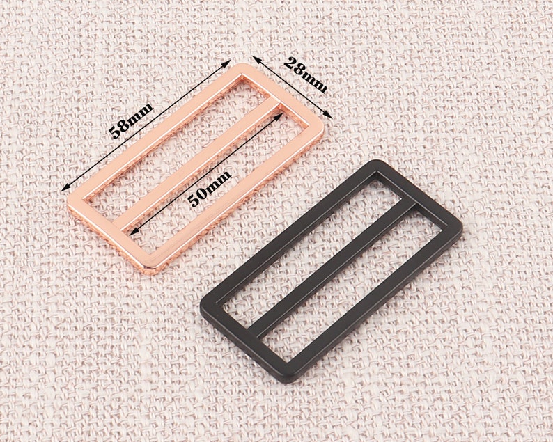 Puede incluir: Dos hebillas deslizantes de metal, una de oro rosa y otra negra. La hebilla de oro rosa mide 28 mm por 58 mm y 50 mm de ancho. La hebilla negra es del mismo tama&ntilde;o.