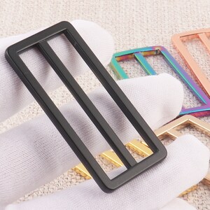 Puede incluir: Un conjunto de cuatro hebillas de cintur&oacute;n de metal en diferentes colores, incluyendo negro, dorado, oro rosa e iridiscente. Las hebillas tienen una forma rectangular con dos barras paralelas en el centro.