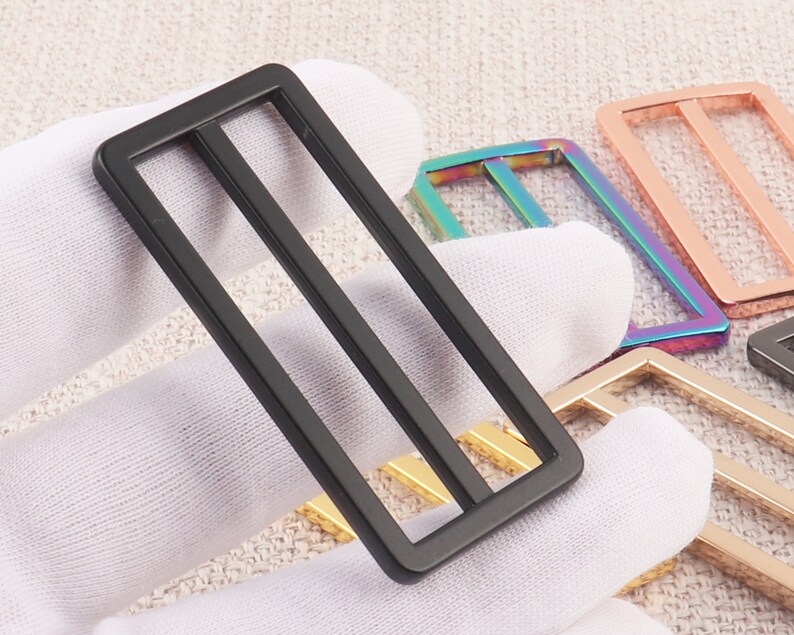 Puede incluir: Un conjunto de cuatro hebillas de cintur&oacute;n de metal en diferentes colores, incluyendo negro, dorado, oro rosa e iridiscente. Las hebillas tienen una forma rectangular con dos barras paralelas en el centro.