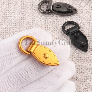 May include: Close-up of several metal bag clasps. One is gold-toned, with a rounded ring and a teardrop-shaped base. The base has a central rivet and decorative stitching. Two other clasps are black, with similar shapes and screw details.