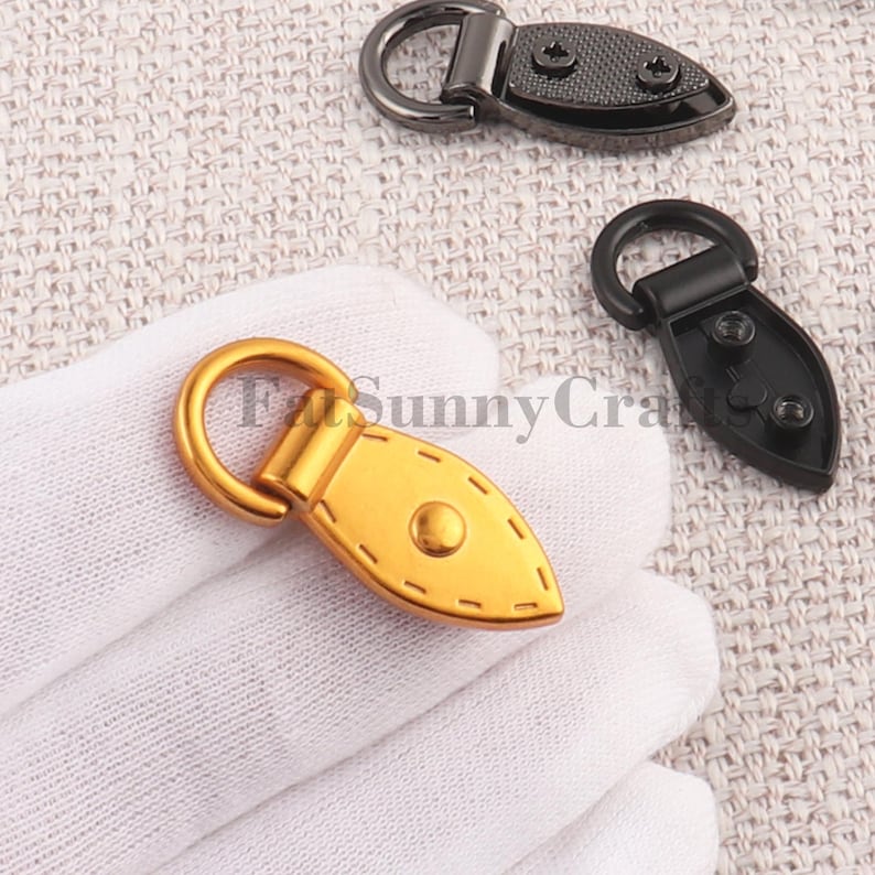 May include: Close-up of several metal bag clasps. One is gold-toned, with a rounded ring and a teardrop-shaped base. The base has a central rivet and decorative stitching. Two other clasps are black, with similar shapes and screw details.