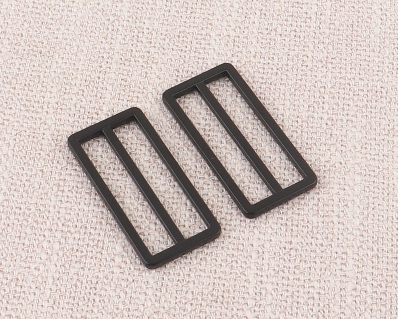 Puede incluir: Dos hebillas deslizantes de metal negro con forma rectangular y dos l&iacute;neas paralelas que atraviesan el centro.