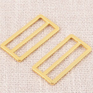 Puede incluir: Dos hebillas deslizantes de metal de color dorado. Las hebillas son rectangulares con dos l&iacute;neas paralelas que atraviesan el centro.