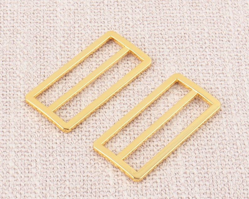 Puede incluir: Dos hebillas deslizantes de metal de color dorado. Las hebillas son rectangulares con dos l&iacute;neas paralelas que atraviesan el centro.