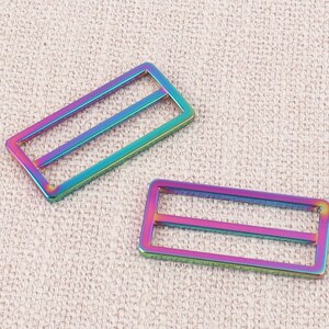 Puede incluir: Dos hebillas deslizantes de metal iridiscente. Las hebillas son rectangulares con dos barras paralelas en el interior. Las hebillas son un arco&iacute;ris de colores, incluyendo rosa, azul, verde y morado.