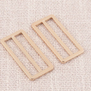 Puede incluir: Dos hebillas deslizantes de metal dorado con forma rectangular y tres l&iacute;neas paralelas.