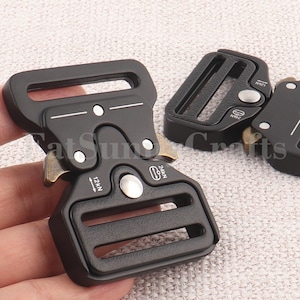 Pode incluir: Close-up de várias fivelas de metal preto. As fivelas têm formato retangular com bordas arredondadas e acabamento fosco. As fivelas têm o texto "12kN" e "24kN" impresso. As fivelas são provavelmente usadas para tiras ou cintos.