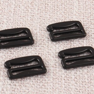May include: Four black plastic bra strap adjusters. The adjusters have two loops and are designed to be used with bra straps.