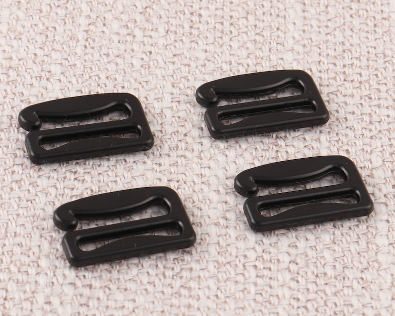May include: Four black plastic bra strap adjusters. The adjusters have two loops and are designed to be used with bra straps.