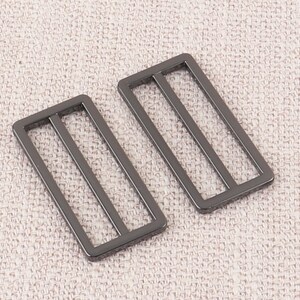 Puede incluir: Dos hebillas deslizantes de metal negro con forma rectangular y tres l&iacute;neas paralelas. Las hebillas est&aacute;n sobre un fondo de tela beige claro.