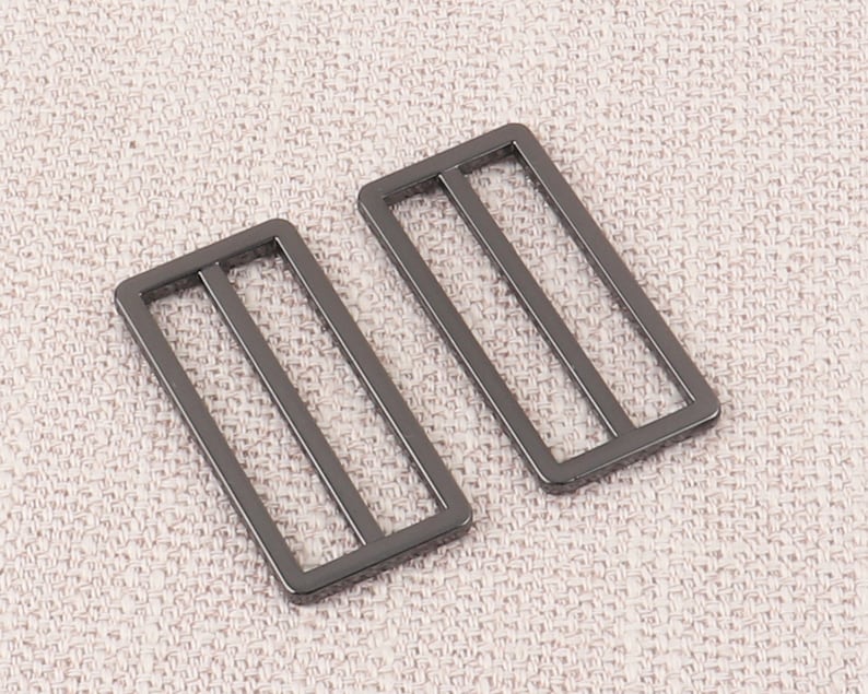 Puede incluir: Dos hebillas deslizantes de metal negro con forma rectangular y tres l&iacute;neas paralelas. Las hebillas est&aacute;n sobre un fondo de tela beige claro.
