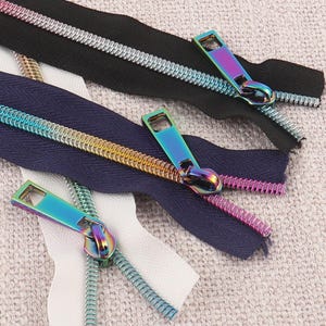 May include: Three zippers with iridescent rainbow-colored metal teeth and matching pulls. One zipper has black fabric tape, one has dark blue fabric tape, and one has white fabric tape. The metallic teeth display a gradient of colors.