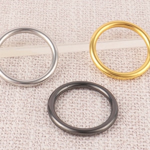 May include: Three metal rings in different finishes: silver, gold, and black. The rings are all the same size and are arranged in a row on a textured fabric background.