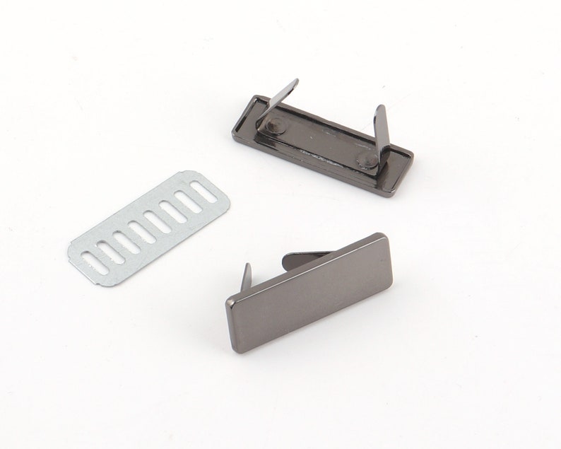 K&ouml;nnte beinhalten: Drei Metall-Geldb&ouml;rsenbeschl&auml;ge. Zwei sind rechteckig mit Stiften, und einer ist eine geschlitzte Metallplatte. Die Beschl&auml;ge haben eine dunkle Gunmetal-Farbe, vor einem wei&szlig;en Hintergrund.