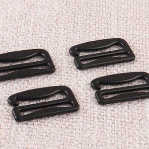 May include: Four black plastic bra strap adjusters. The adjusters have two loops and are designed to be used with bra straps.