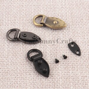 May include: Close-up of three metal toggle clasps in antique brass and black finishes. Each clasp features a D-ring and a decorative oval plate with screw holes. Includes screws and a small plate.