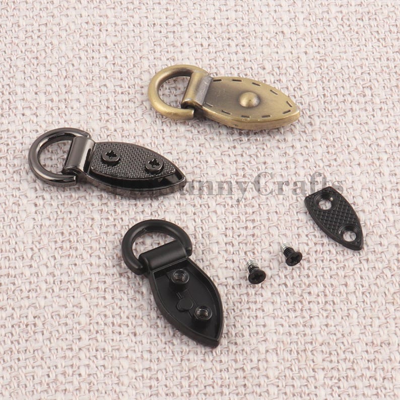 May include: Close-up of three metal toggle clasps in antique brass and black finishes. Each clasp features a D-ring and a decorative oval plate with screw holes. Includes screws and a small plate.