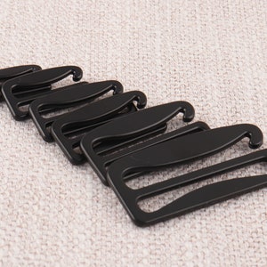 May include: Five black plastic bra strap adjusters. The adjusters have a hook on one end and a slider on the other end.
