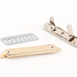 May include: A set of three metal hardware pieces for attaching a strap or handle to a bag or other project. The pieces are silver, gold, and a silver plate with holes.