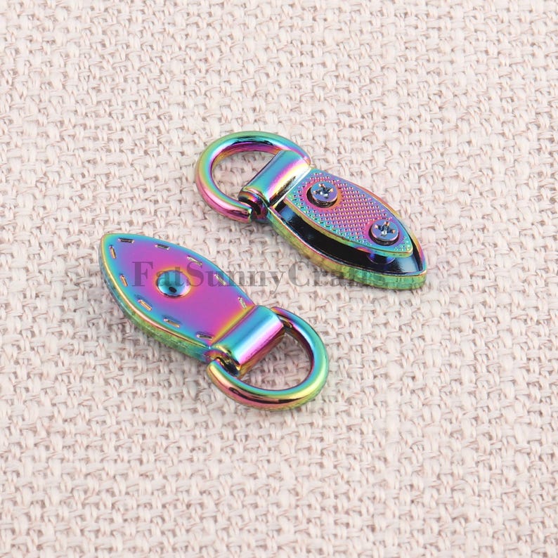 May include: Two iridescent metal purse clasps with a rainbow finish. Each clasp has a rounded loop and a teardrop-shaped body secured with screws. The clasps are on a textured, beige surface.