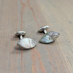 May include: A pair of silver cufflinks with oval-shaped abalone shell inlays. The iridescent shells display a mix of white, gray, and blue hues. The cufflinks are set against a wooden surface.