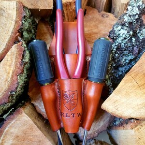 May include: A brown leather tool holder with a shield emblem and the letters "RLTW" embossed on the front. It holds red and blue pliers and two screwdrivers with black handles. The tools are displayed against a backdrop of stacked firewood.