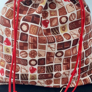 Könnte beinhalten: Eine weiße Kordeltasche mit einem Schokoladenriegelmuster. Die Tasche hat rote Kordeln mit Quasten. Die Tasche liegt auf einer schwarzen Oberfläche mit Holzspießen.