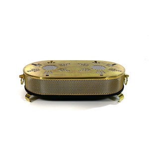 Op de afbeelding: Een ovale, goudkleurige metalen chafing dish met een gaasbodem en decoratieve uitsnijdingen. De schaal heeft vier kleine pootjes en twee handvatten.