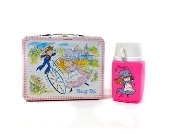 May include: A pink and white lunchbox with a checkered pattern and a picture of a girl and boy riding a bicycle. The lunchbox says "Polly Pal" on the front. A matching pink and white flask with a picture of a girl holding a basket is also included.