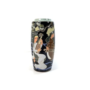 May include: A black ceramic vase with a green top, featuring a painted scene of two women playing music in a landscape. The vase has a cylindrical shape and is decorated with intricate details.