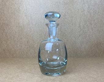 Pode incluir: Uma garrafa de vidro transparente com uma rolha de vidro azul claro. A garrafa tem um corpo arredondado e uma base larga.