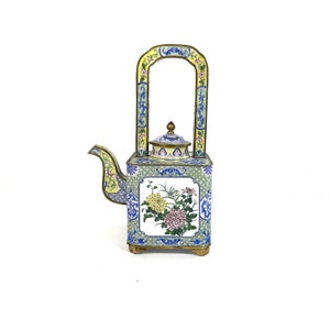 May include: A decorative, square-shaped teapot with a handle, spout, and lid. The teapot features a floral design with yellow, pink, and green flowers on a white background. The handle and spout are yellow and blue, and the base is gold.