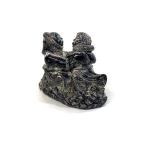 May include: A black soapstone sculpture of two people dancing. The sculpture is carved in a traditional Inuit style and depicts a man and a woman in traditional clothing.