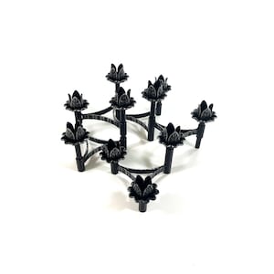 May include: A black metal candle holder with nine floral-shaped candle cups. The candle holder has a geometric design with curved arms connecting the candle cups. The piece is made of metal and sits on a white surface.