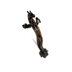 May include: Bronze-coloured door handle shaped like a rearing horse. The handle has detailed features, including the horse's mane, tail, and hooves. The horse is on its hind legs, with front legs raised. The handle is likely made of metal.