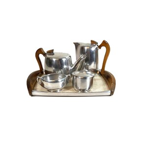 Op de afbeelding: Een theeservies van zilverkleurig metaal bestaande uit een theepot, een melkkannetje, een suikerpot en een kan, allemaal voorzien van donkerbruine houten handvatten. De set staat op een rechthoekig dienblad met bijpassende houten zijhandvatten.