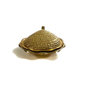 May include: A gold-colored, seashell-shaped trinket box with a textured surface. The box has a lid with a small handle and a scalloped edge. The box is made of metal and sits on a white surface.