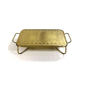 May include: A rectangular brass serving tray with a perforated surface and curved handles. The tray is supported by a delicate, angled frame. The brass has a warm, golden hue, and the design is simple and elegant.