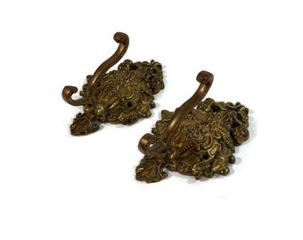 May include: Two antique brass wall hooks with ornate floral and scrollwork detailing. The hooks have a curved design and are mounted on decorative backplates. The brass finish has a warm, aged patina, adding to their vintage appeal.