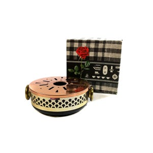 May include: A copper tea warmer with a perforated top and brass handles, alongside a black and white checked box adorned with a red rose and illustrations. The warmer features a black base and a decorative patterned band.