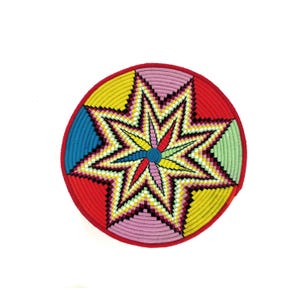 May include: A circular woven basket with a colourful starburst pattern. The basket has a red border and a central star design in yellow, pink, blue, green, and white. The star's centre is a small, blue circle.