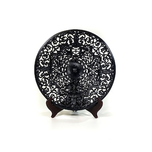 Peut inclure: Décoration murale ronde en métal noir avec un motif de filigrane complexe. La suspension est ronde et présente un motif floral central. Elle est présentée sur un support en bois.